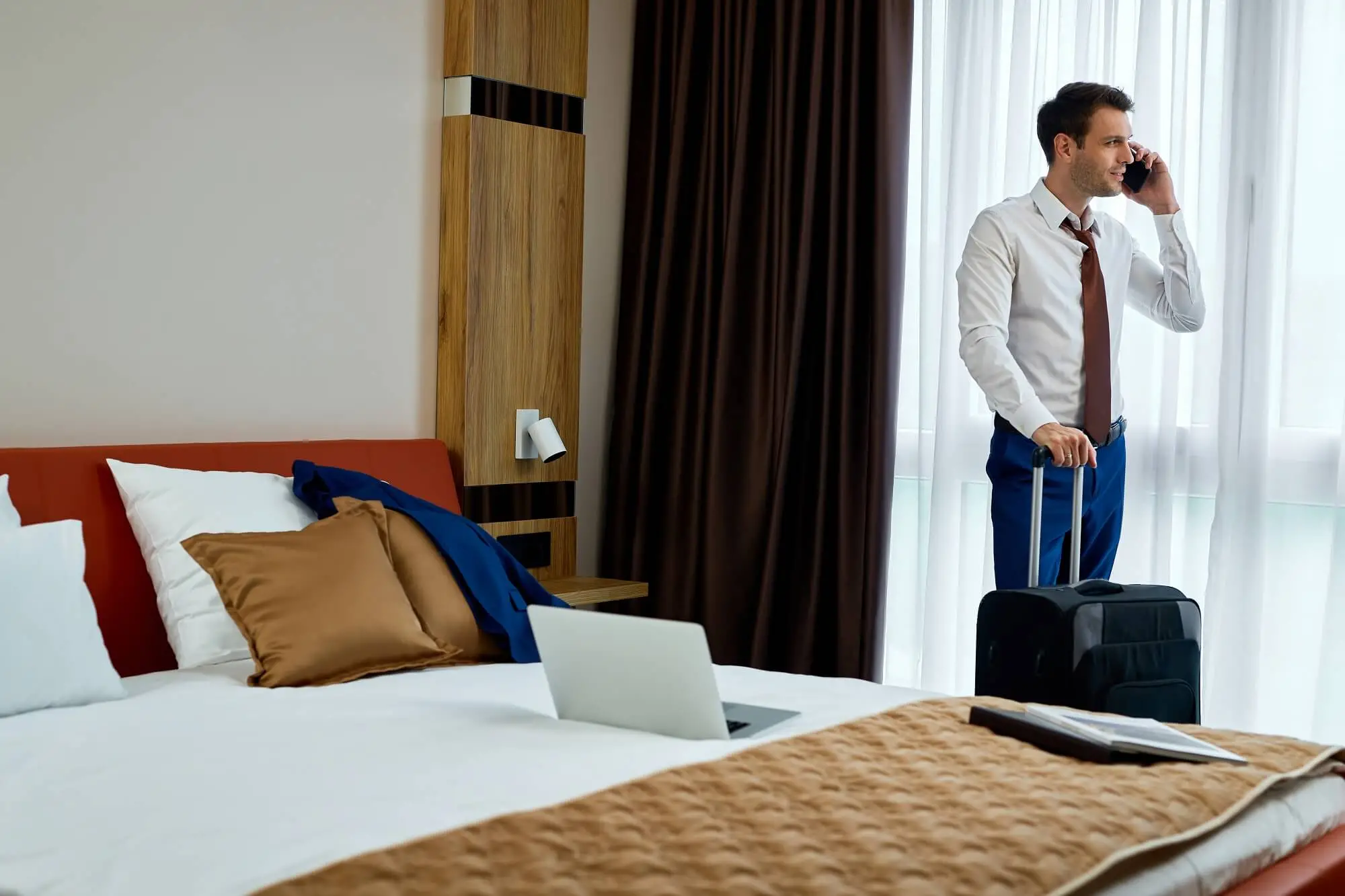 smiling-businessman-communicating-mobile-phone-while-standing-with-travel-bag-hotel-room